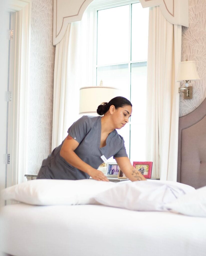 Vella cleaner making a bed and arranging pillows in a bright, tidy apartment bedroom.