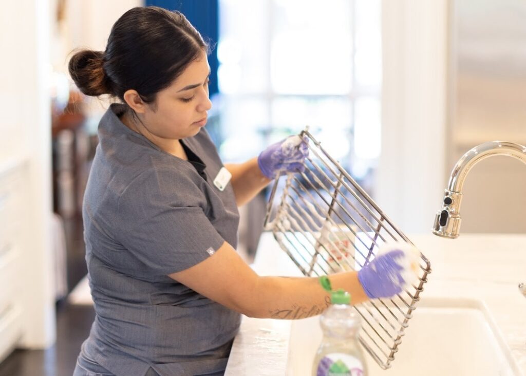 This image shows a person cleaning a metal dish rack at a sink This image shows a person cleaning a metal dish rack at a sink. The individual is wearing gloves and working on a kitchen countertop