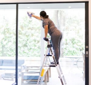A professional cleaner wiping a large glass window while standing on a ladder.