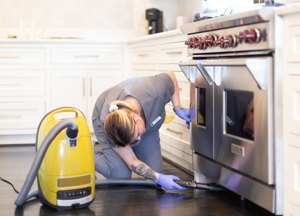 A professional cleaner using a vacuum to clean under a kitchen stove in a modern kitchen A professional cleaner using a vacuum to clean under a kitchen stove in a modern kitchen; remember to clean your sink thoroughly for a spotless kitchen.