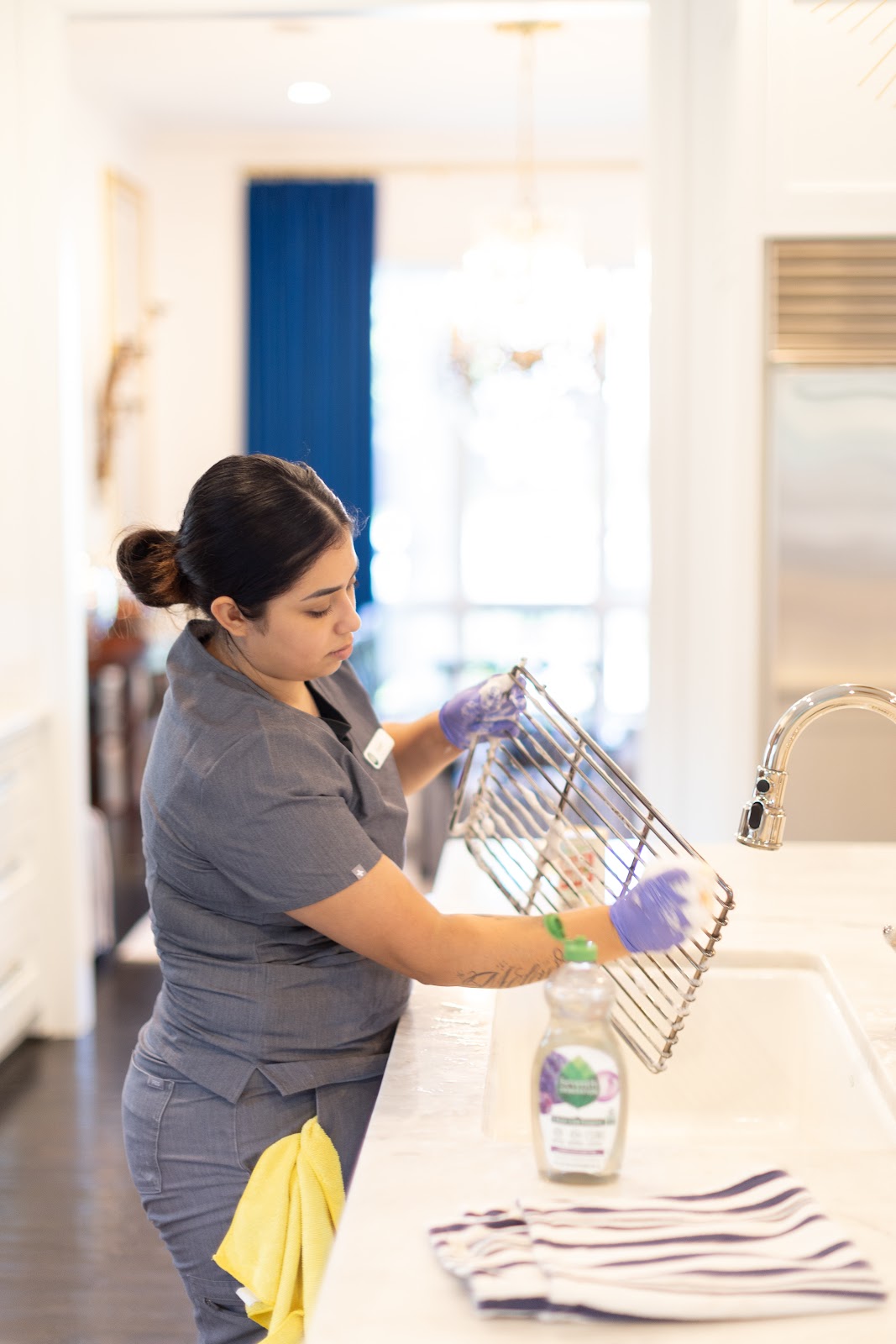 Housekeeping staff member cleaning a metal kitchen rack Housekeeping staff member cleaning a metal kitchen rack at a sink in a well-lit modern kitchen, wearing gloves and a uniform.