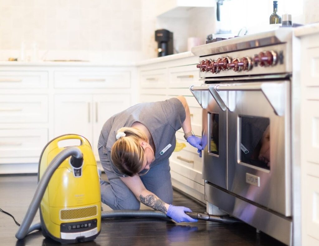 a person cleaning the kitchen of a home. This image depicts a person cleaning the kitchen of a home. They are kneeling on the floor, wearing gloves, and using a vacuum cleaner to clean under the stove.
