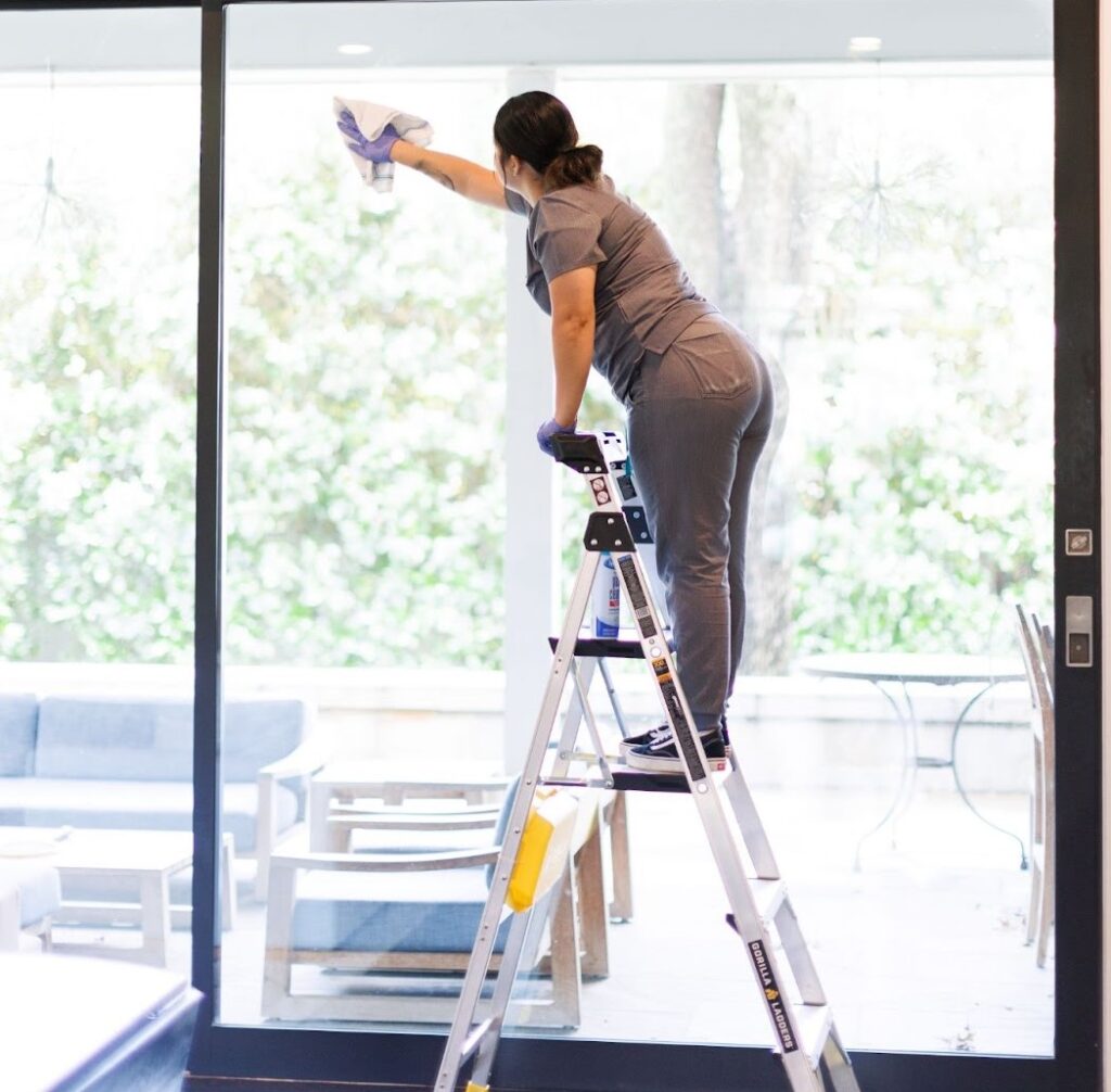 Woman standing on the stepladder cleaning the window A woman is standing on a stepladder, cleaning a large window in a home. She's wearing gloves and holding a cleaning cloth.