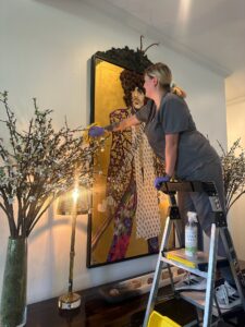 Vella cleaner standing on a ladder dusting artwork and shelves with eco-friendly products during a Dallas deep clean.