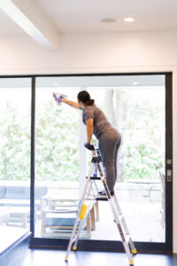 A professional Vella cleaner standing on a ladder, wiping a large glass door inside a bright, modern home.