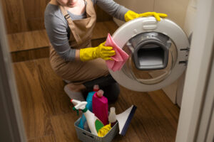A Vella cleaner in gloves wipes a washing machine door with a cloth, surrounded by eco-friendly cleaning supplies.