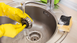 Hands wearing yellow rubber gloves cleaning a sink strainer under running water in a stainless steel kitchen sink.