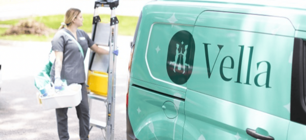 Housekeeping professional with cleaning supplies beside a Vella service van. A housekeeping professional stands by a teal van labeled "Vella," carrying cleaning supplies and a ladder, ready for service.