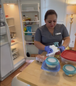 Housekeeper in uniform wiping a food container while cleaning and organizing a refrigerator in a home kitchen.