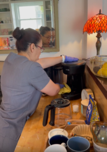 Housekeeper in uniform cleaning a black Mr. Coffee machine on a wooden kitchen counter with gloves on.